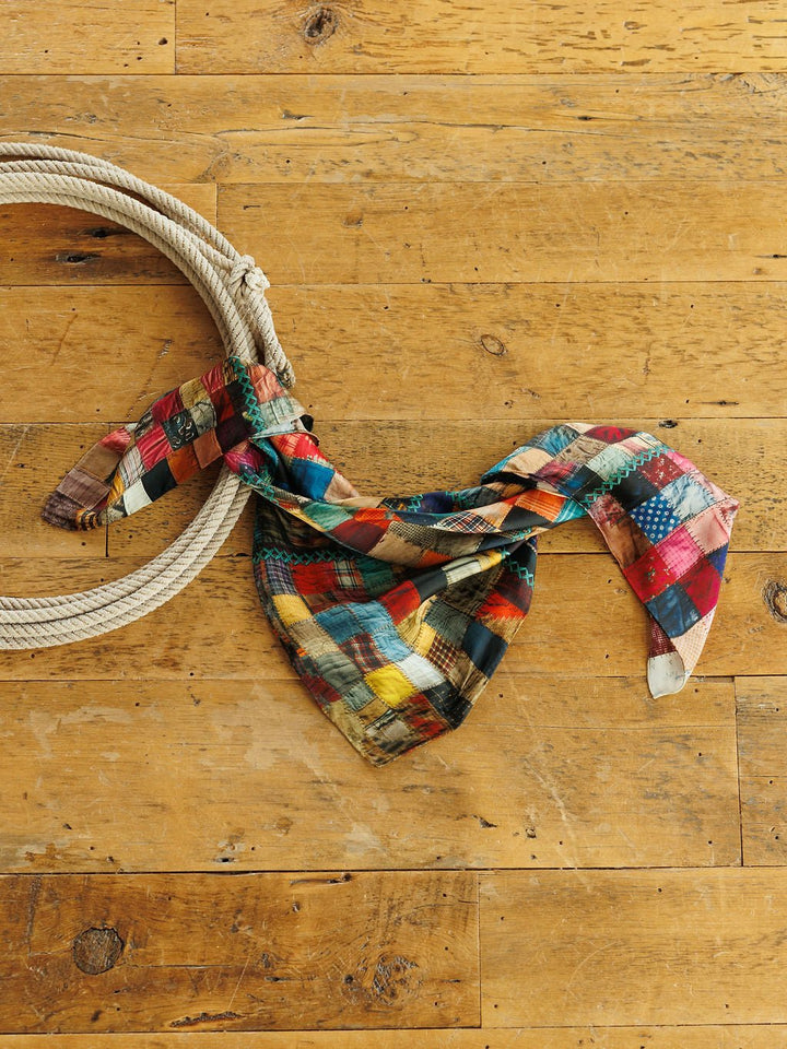 A colorful Quilt Silk Scarf from Savannah Sevens western life&style draped over a coiled rope on a wooden floor.