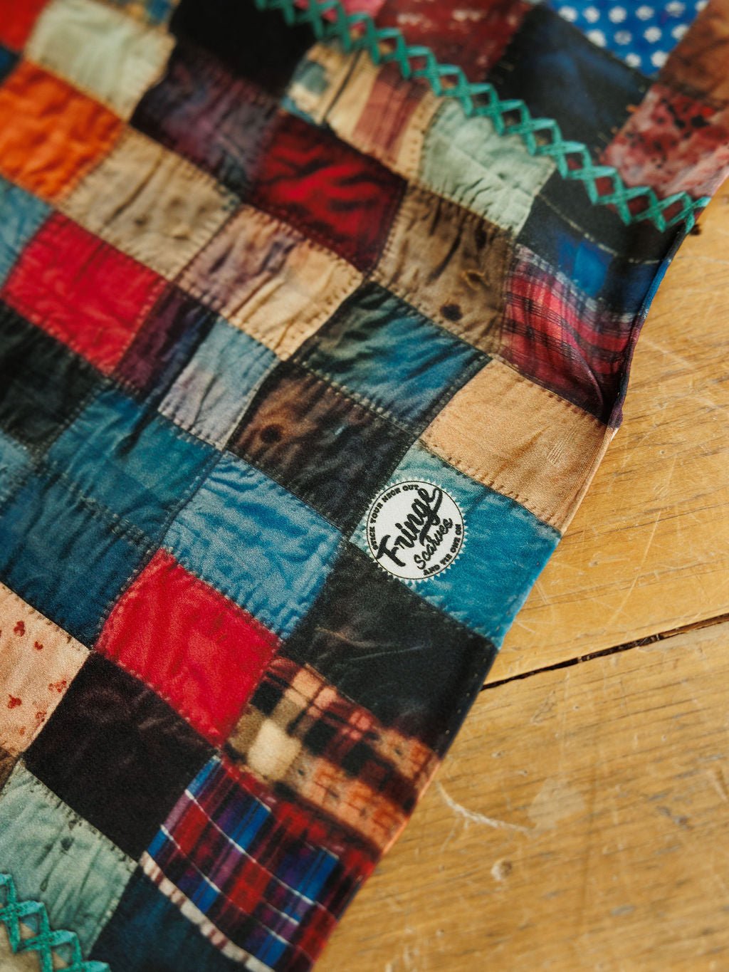 A Quilt Silk Scarf by Savannah Sevens western life&style featuring a multicolored patchwork pattern and a circular brand logo near the edge.