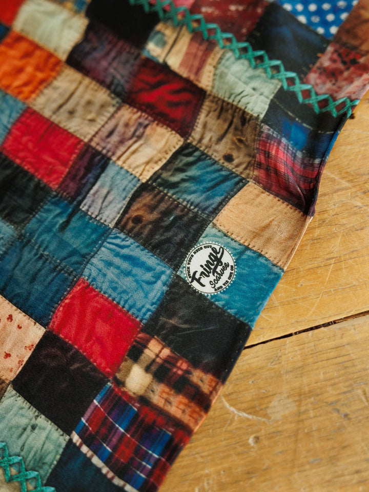 A Quilt Silk Scarf by Savannah Sevens western life&style featuring a multicolored patchwork pattern and a circular brand logo near the edge.