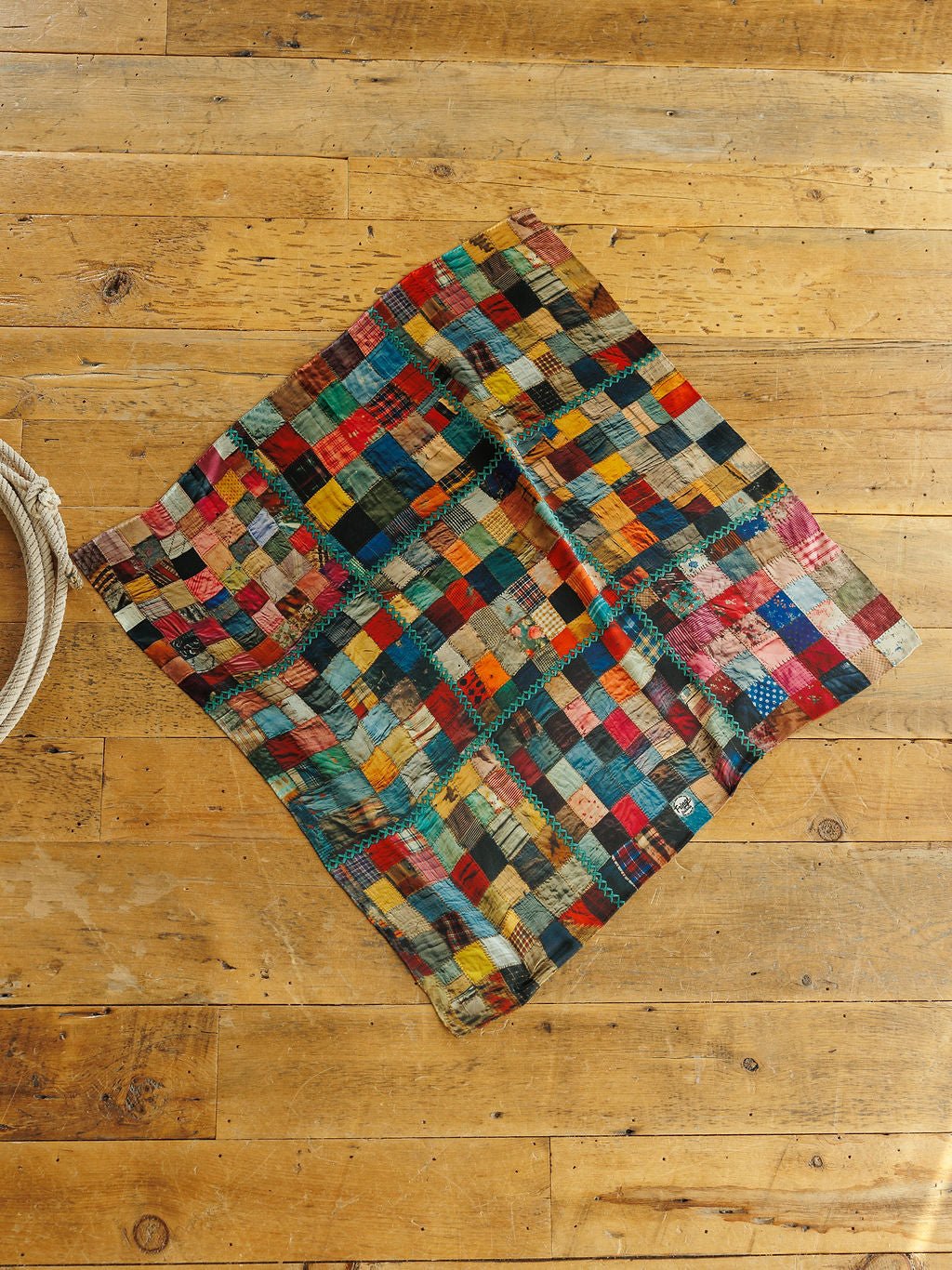 Savannah Sevens western life&style Quilt Silk Scarf featuring a multicolored patchwork of squares with teal zigzag stitching, laid flat on a wooden floor.
