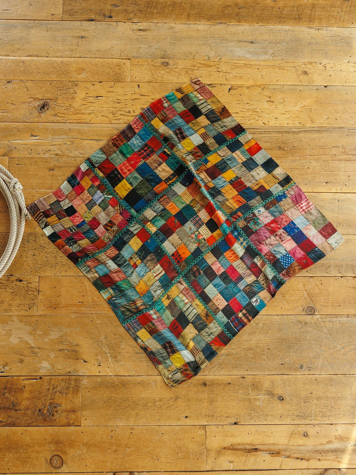 Savannah Sevens western life&style Quilt Silk Scarf featuring a multicolored patchwork of squares with teal zigzag stitching, laid flat on a wooden floor.
