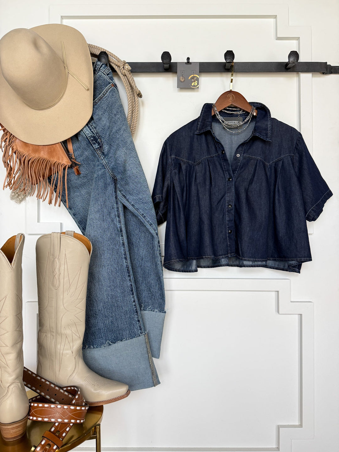 The Quinby Denim Tencel Pearl Snap jeans by Savannah Sevens western life{&}style and a short-sleeve oversized denim shirt hang on hooks beside a beige hat, fringed scarf, and cream cowboy boots with a studded brown belt, all arranged against a white wall.