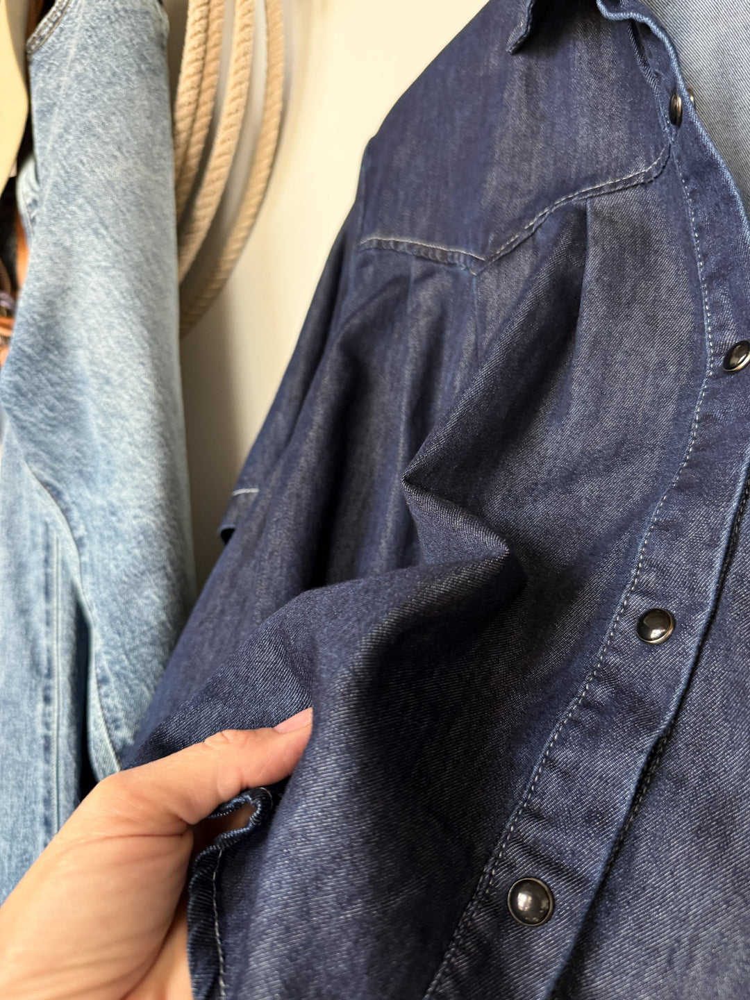A hand is holding and gently pulling the fabric of the Quinby Denim Tencel Pearl Snap shirt by Savannah Sevens western life{&}style, while another light blue denim garment and coiled rope are visible in the background.