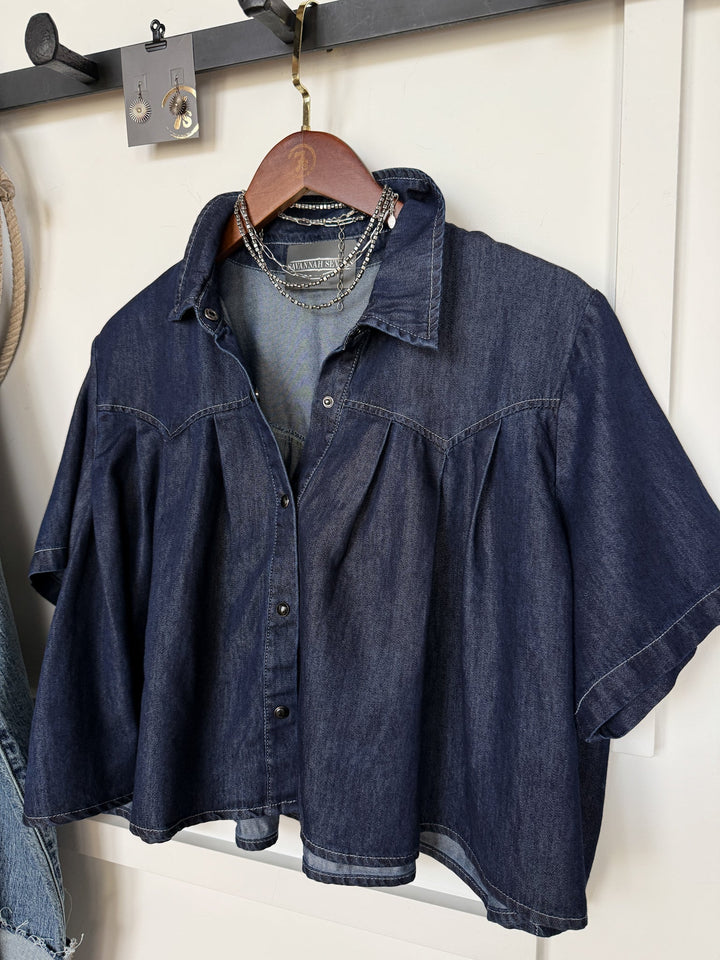 The Quinby Denim Tencel Pearl Snap shirt by Savannah Sevens western life{&}style, featuring an oversized fit in dark denim with short sleeves and pearl snap buttons, is displayed on a hanger layered with silver chain necklaces and hanging on a hook against a white wall.