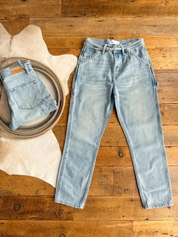 Light blue non-stretch denim jeans are laid flat on a wooden floor. Next to them, Savannah Sevens' The Quinn High Rise 90's Straight Carpenter Jean is folded neatly on a round tray atop a cream-colored hide rug.