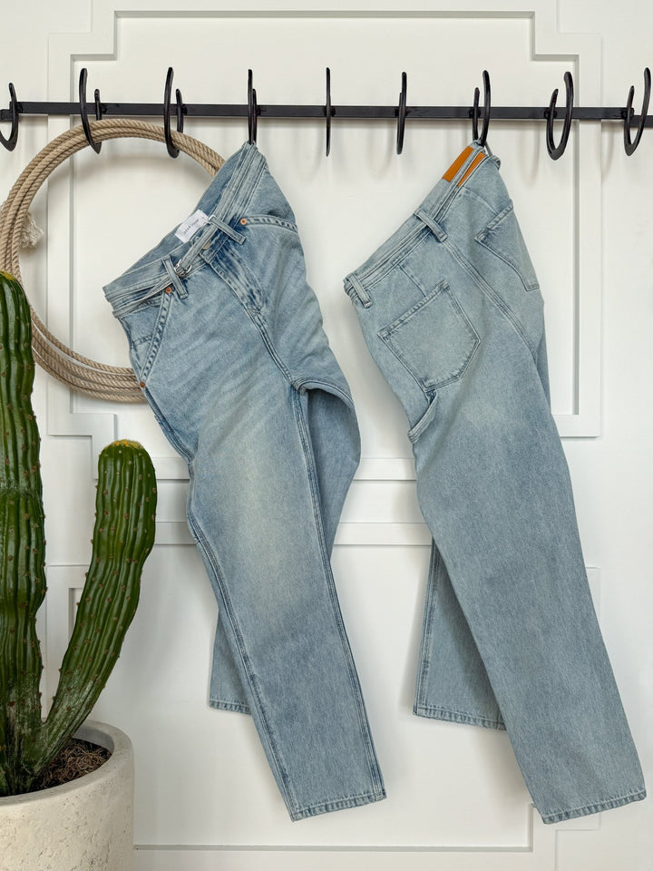 Two pairs of Savannah Sevens western life{&}style The Quinn High Rise 90's Straight Carpenter Jeans hang side by side on black hooks against a white paneled wall, with a tall green cactus in a white pot to the left and a coiled rope in the background.
