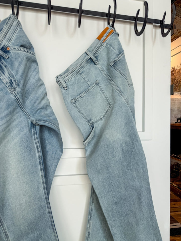 Two pairs of The Quinn High Rise 90's Straight Carpenter Jeans by Savannah Sevens western life{&}style in light blue hang on black hooks against a white wall, with part of a room and decor seen in the background.