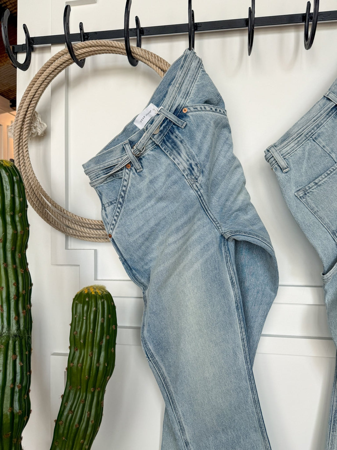 A pair of light blue The Quinn High Rise 90's Straight Carpenter Jeans by Savannah Sevens western life{&}style hang on a black rack, with a large rope circle and two tall green cacti nearby against a white wall.