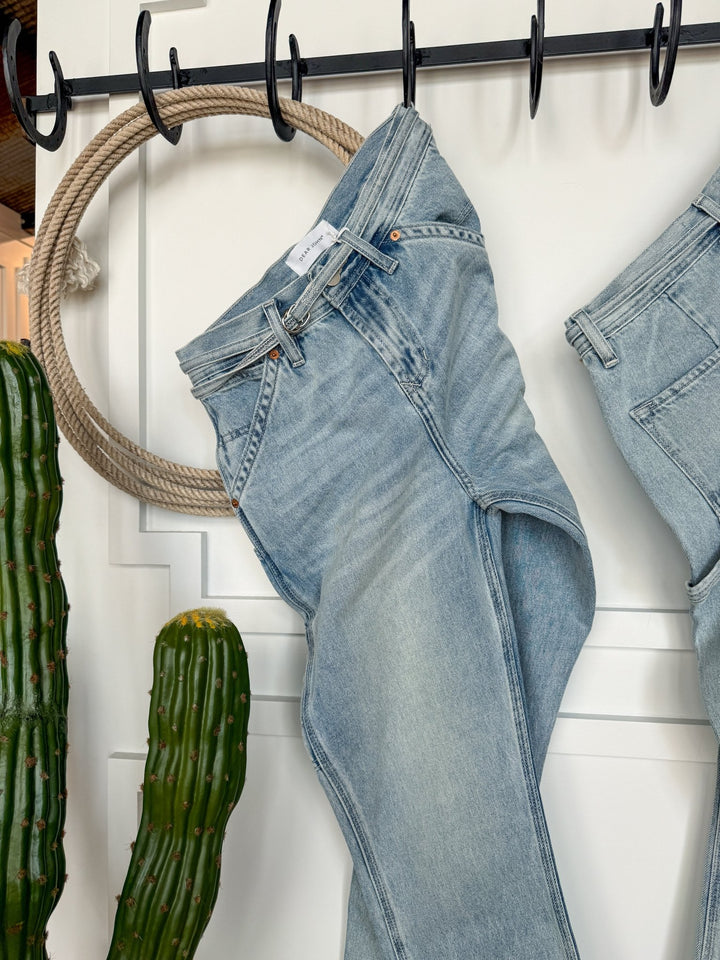 A pair of light blue The Quinn High Rise 90's Straight Carpenter Jeans by Savannah Sevens western life{&}style hang on a black rack, with a large rope circle and two tall green cacti nearby against a white wall.