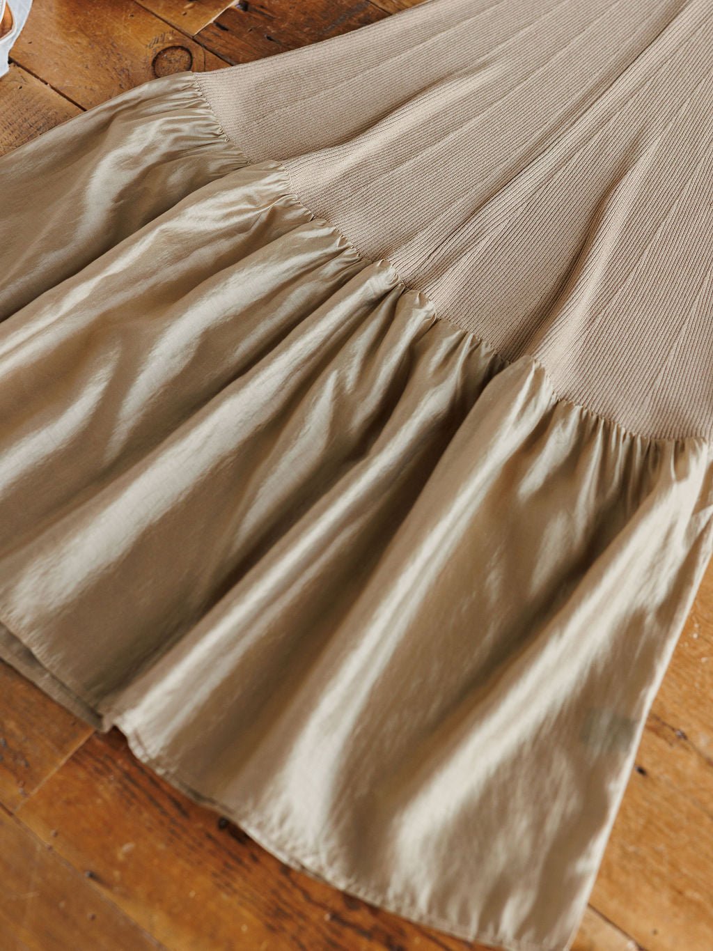 The Quivera Bronze Champagne Ribbed Dress by Savannah Sevens western life{&}style showcases a ribbed, stretchy top and a shiny, satin-like flared skirt with a subtle ruffle hem, displayed on a wooden surface.
