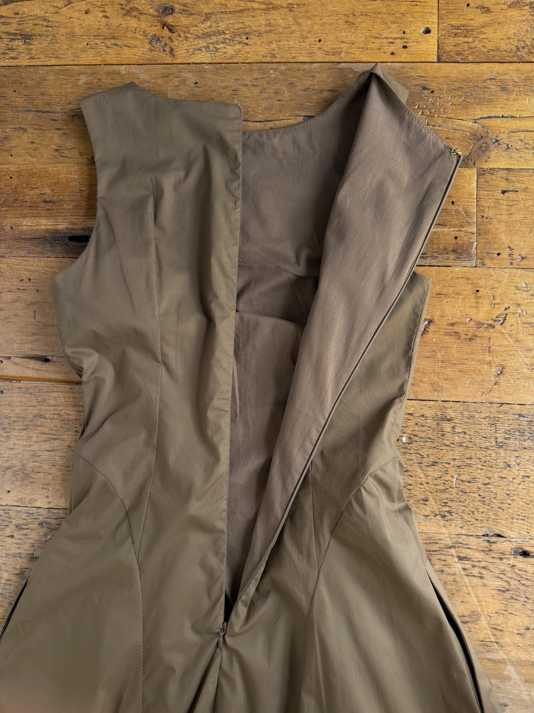 The Ralston Mushroom Sleeveless Dress by Savannah Sevens western life{&}style, an olive green cotton A-line dress, is displayed flat on a wooden surface with its back partially unzipped to reveal the brown inner lining.
