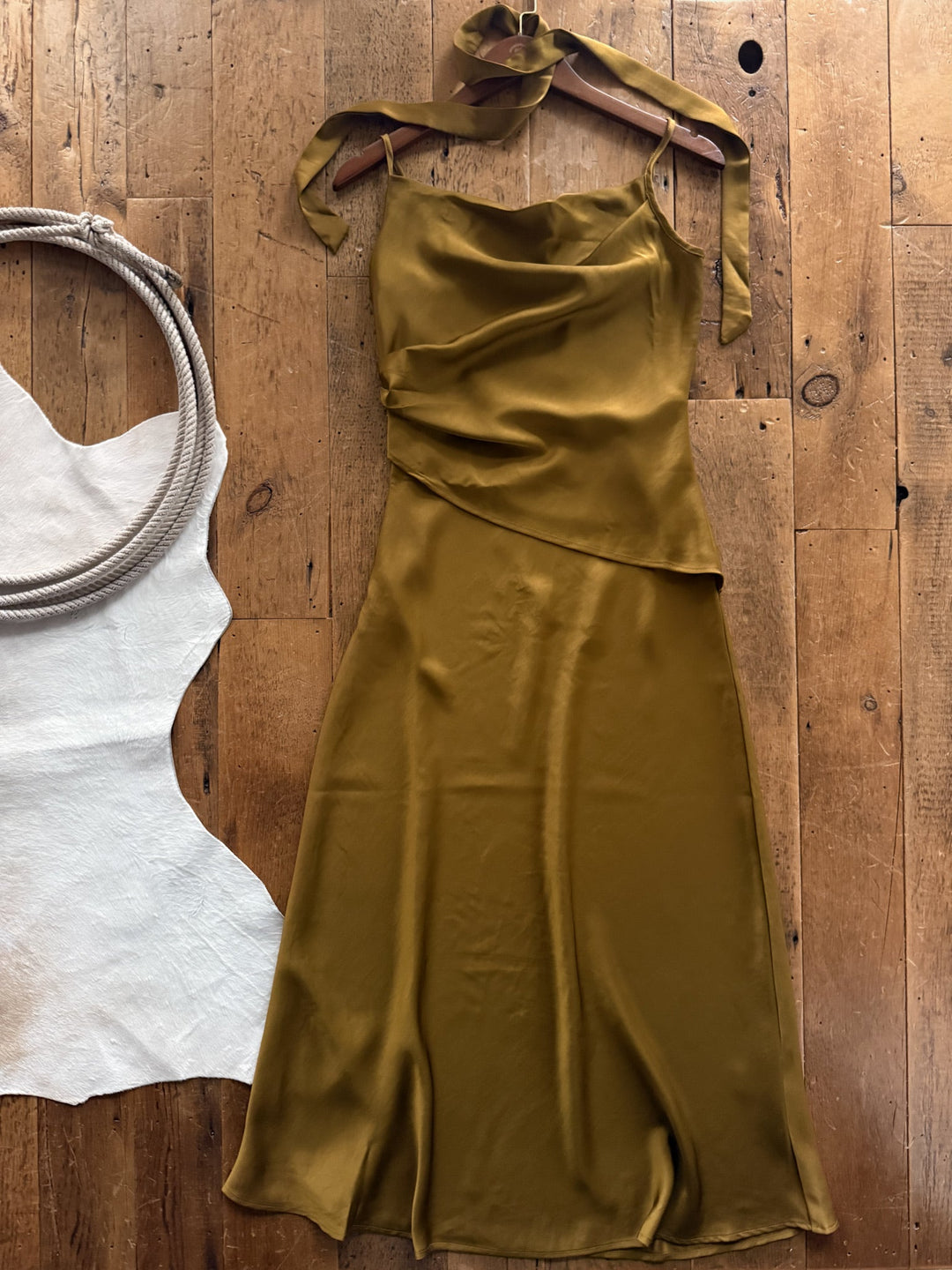 Ramona Olive Chartreuse Sleeveless Dress in olive green satin with a cowl neckline and halter tie, displayed on a hanger next to a coiled rope and a white cowhide rug.