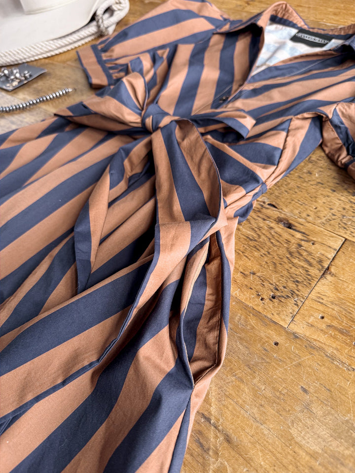 The Ramona Striped Puff Sleeve Dress by Savannah Sevens western life{&}style, featuring brown and navy stripes, puff sleeves, a tied waist, is displayed on a wooden surface alongside partial glimpses of a white bag and jewelry.