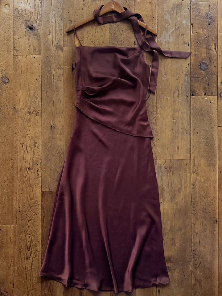 Ramona Wine Sleeveless Dress in deep burgundy, featuring a cowl neckline and tie-detail shoulder strap, hanging as a fitted satin midi dress on a wooden hanger against a rustic wood floor.