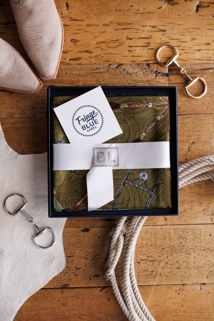A Ranch Gear Wild Rag by Savannah Sevens western life{&}style, presented in a gift box with white ribbon and luxury blue label, sits on a wooden table beside tan boots, cowhide, rope coil, and horse bit accessories.