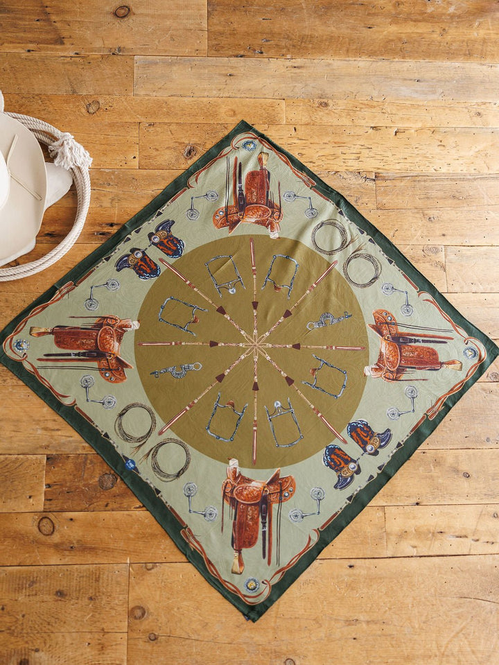 The Ranch Gear Wild Rag by Savannah Sevens western life{&}style is a square jacquard silk scarf with a green and beige Western pattern—saddles, boots, spurs—styled on rustic wood with a white hat and coiled rope. Perfect as a western wild rag.