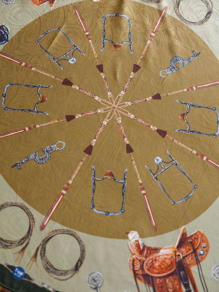 The Ranch Gear Wild Rag by Savannah Sevens western life{&}style features a jacquard silk scarf with a bold golden circle, ten brown and red spears, ornate bridles, and saddles along the edges for a luxurious touch.