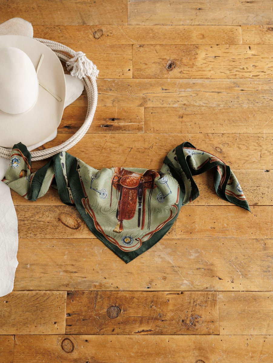 The Ranch Gear Wild Rag by Savannah Sevens western life{&}style, featuring a green and brown jacquard pattern, is displayed on a wooden floor beside a coiled rope and beige cowboy hat for a luxe western look.