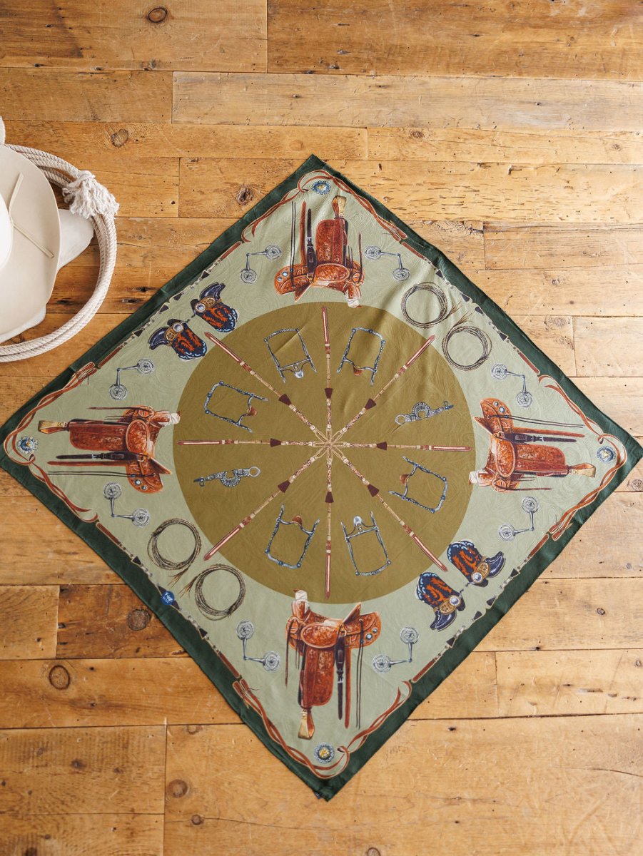 The Ranch Gear Wild Rag by Savannah Sevens western life{&}style is a square jacquard silk scarf with a green and beige Western pattern—saddles, boots, spurs—styled on rustic wood with a white hat and coiled rope. Perfect as a western wild rag.