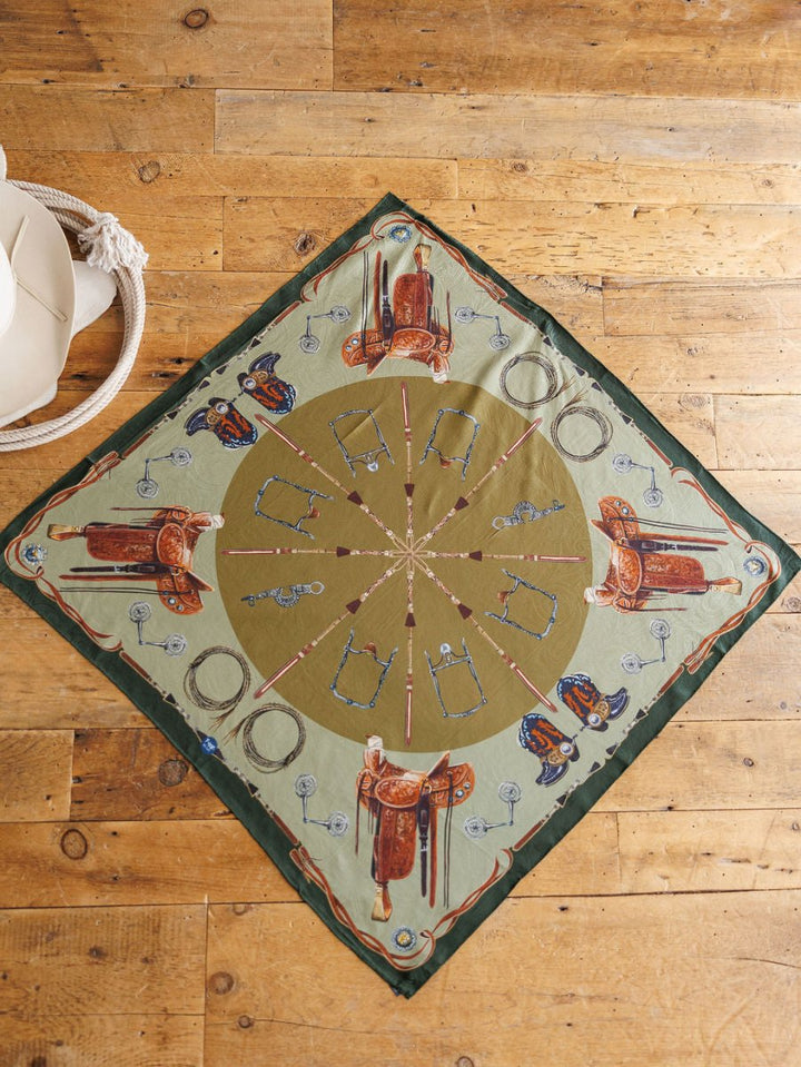 The Ranch Gear Wild Rag by Savannah Sevens western life{&}style is a square jacquard silk scarf with a green and beige Western pattern—saddles, boots, spurs—styled on rustic wood with a white hat and coiled rope. Perfect as a western wild rag.