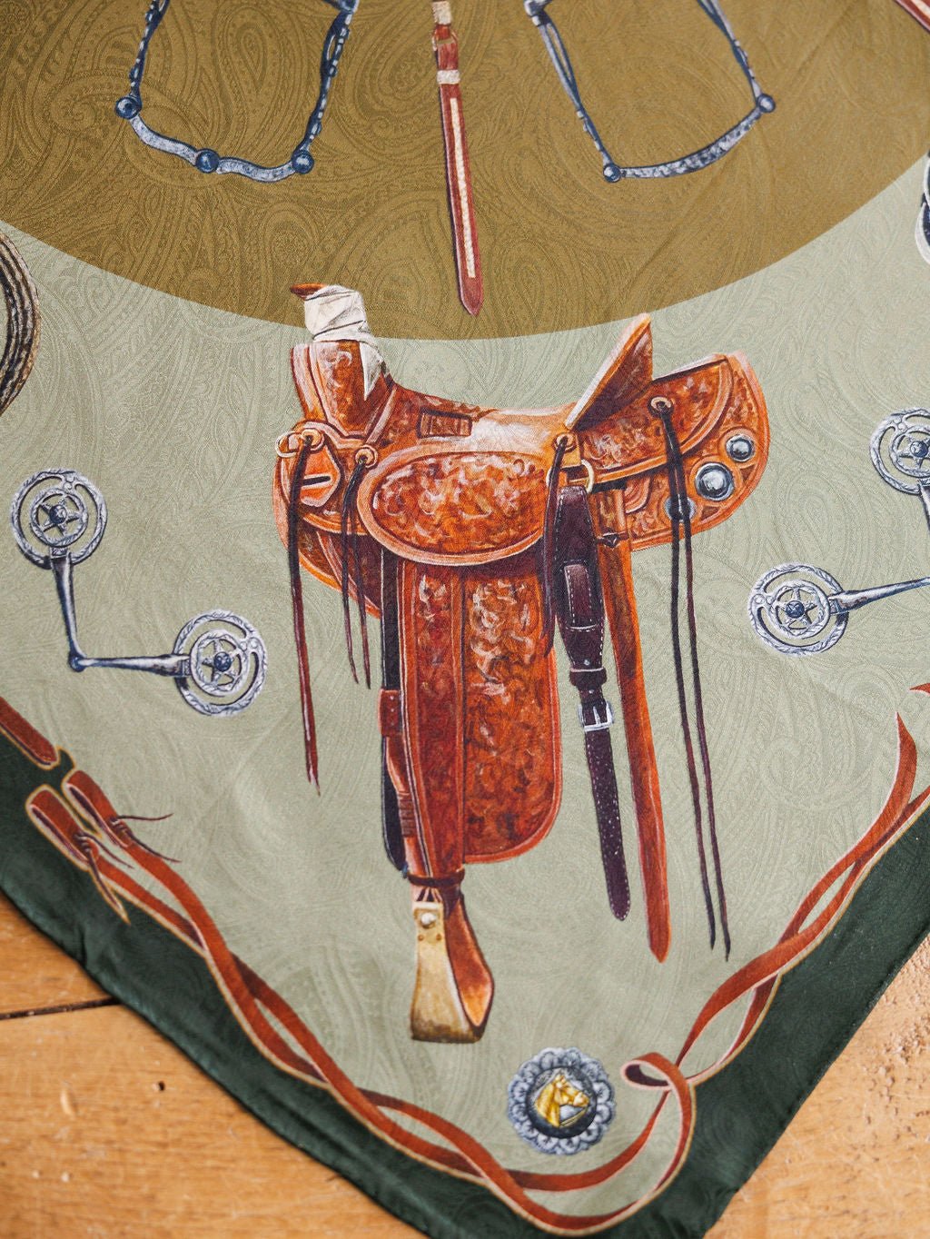 The Ranch Gear Wild Rag by Savannah Sevens western life{&}style features a brown leather Western saddle with silver accents, set on a jacquard silk scarf background bordered by bits and spurs atop wood.