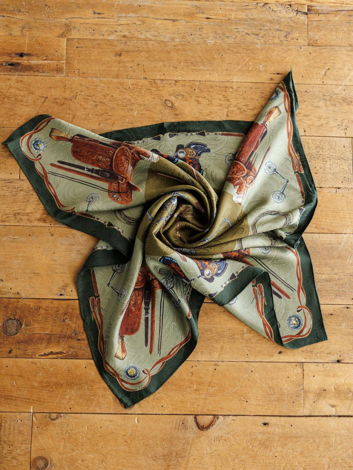 The Savannah Sevens western life{&}style Ranch Gear Wild Rag, a green jacquard silk scarf with an equestrian-themed print of saddles, riding crops, and horse bits, is loosely twisted on a wooden floor, showcasing its luxury blue label design.
