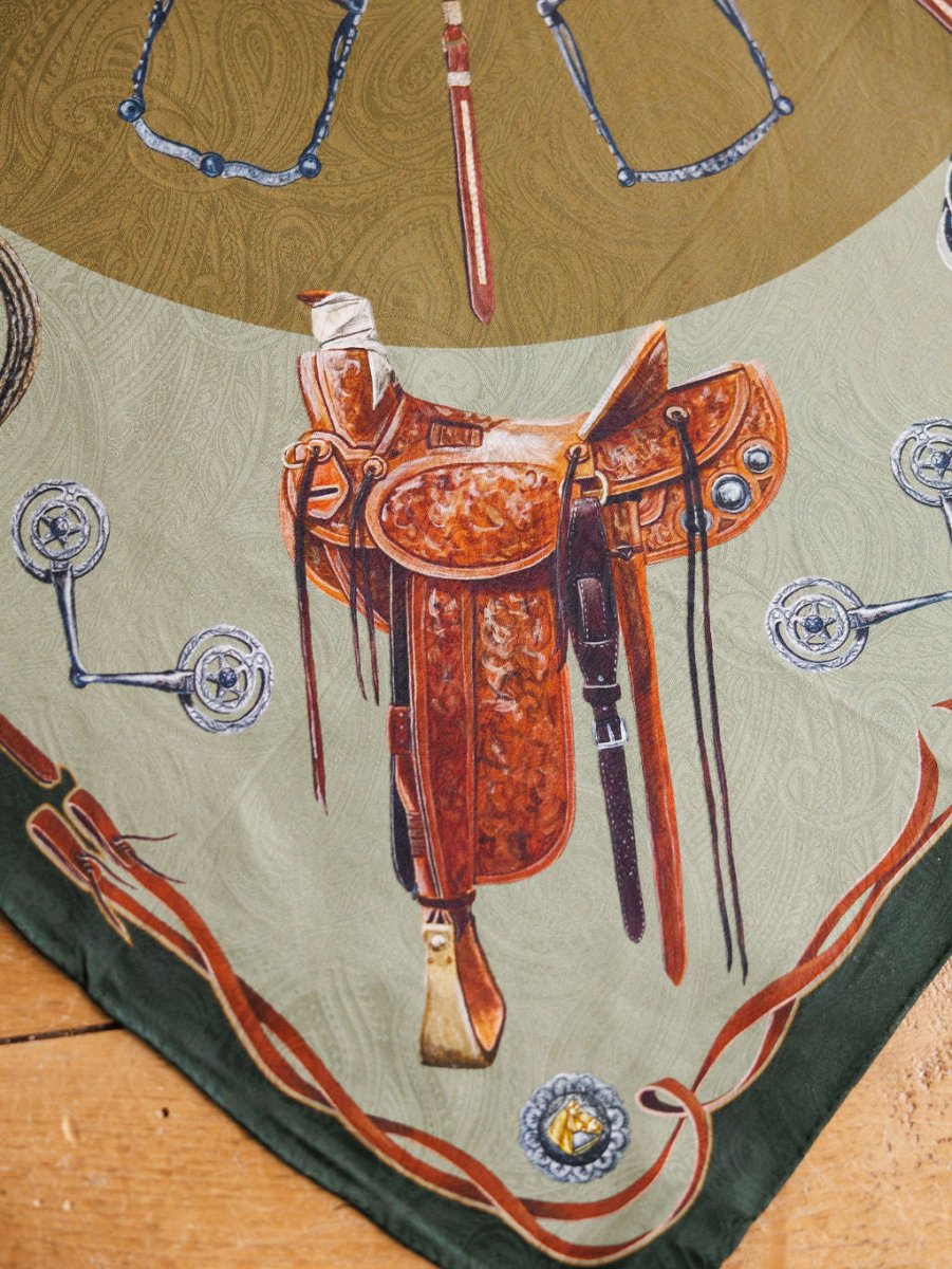 The Ranch Gear Wild Rag by Savannah Sevens western life{&}style features a brown leather Western saddle with silver accents, set on a jacquard silk scarf background bordered by bits and spurs atop wood.