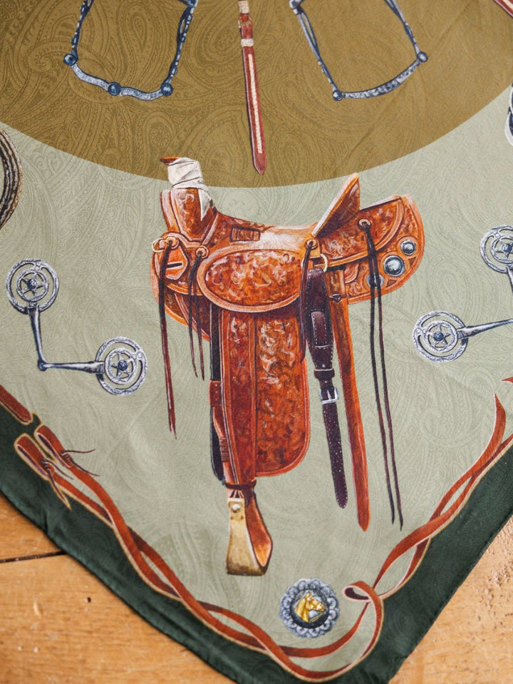The Ranch Gear Wild Rag by Savannah Sevens western life{&}style features a brown leather Western saddle with silver accents, set on a jacquard silk scarf background bordered by bits and spurs atop wood.