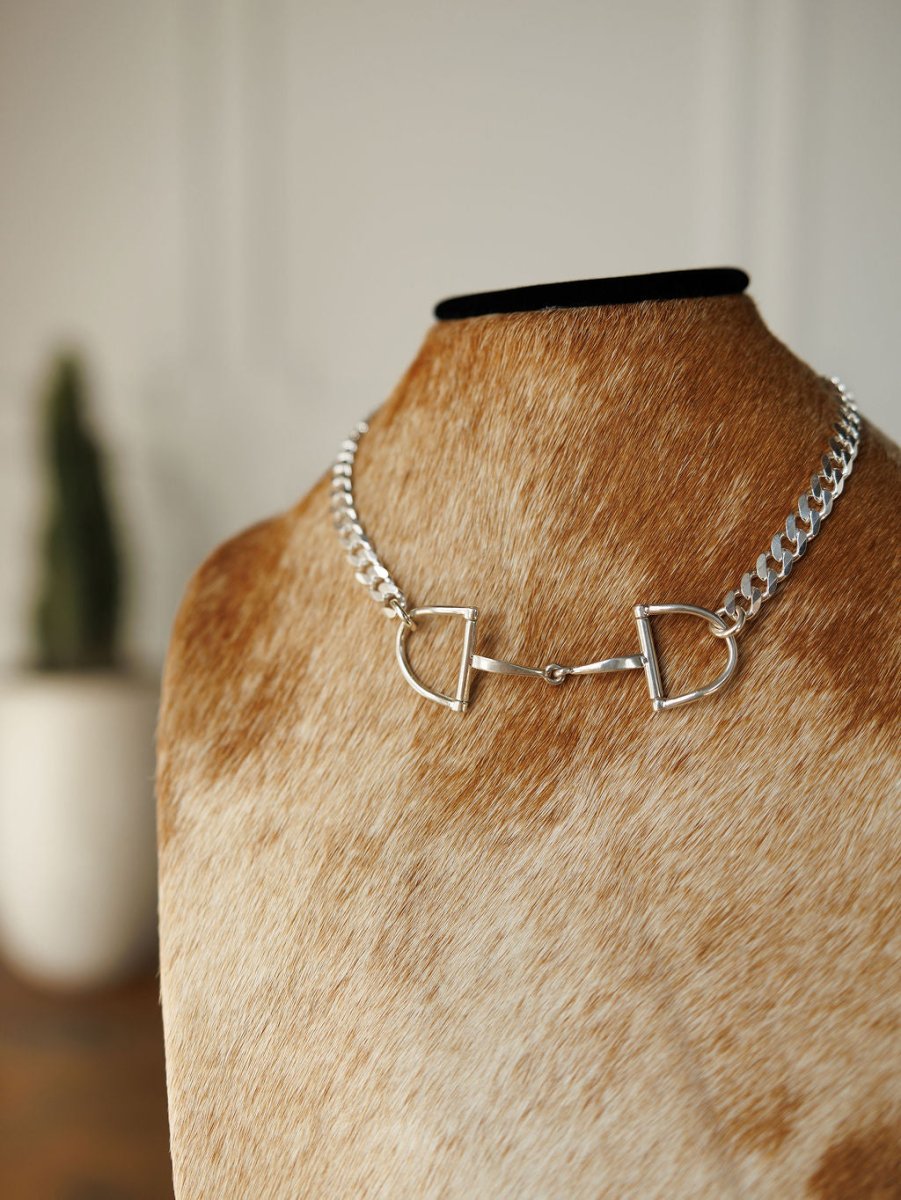 The Ranchero Necklace by Savannah Sevens western life{&}style, featuring a sterling silver snaffle bit and curb link chain, is shown on a brown and white cowhide bust with a potted plant and white wall in the background.