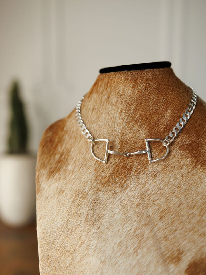 The Ranchero Necklace by Savannah Sevens western life{&}style, featuring a sterling silver snaffle bit and curb link chain, is shown on a brown and white cowhide bust with a potted plant and white wall in the background.