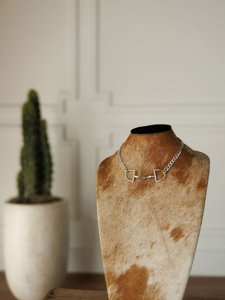The Ranchero Necklace by Savannah Sevens western life{&}style, a 925 Italy sterling silver snaffle bit on a curb link chain, is shown on a cowhide bust with a blurred potted cactus in front of a white paneled wall.