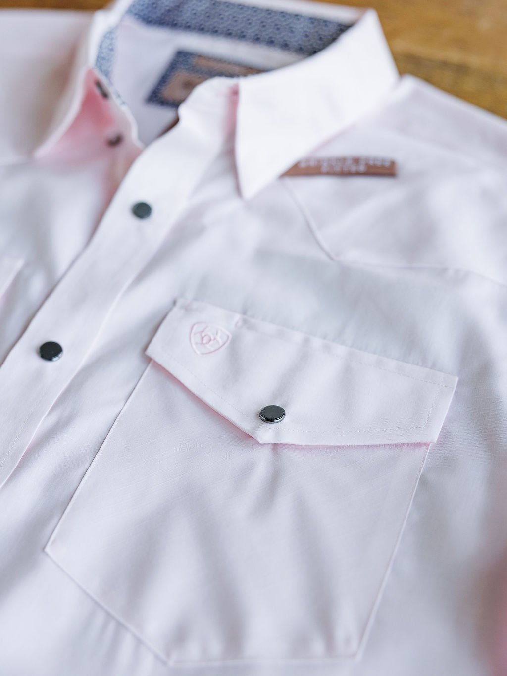 A close-up of the Randall Ariat Men's Light Pink Button Up by Savannah Sevens western life{&}style, made from wrinkle-free cotton with black snap buttons, a front pocket with an embroidered logo, and a patterned inner collar laid on wood.
