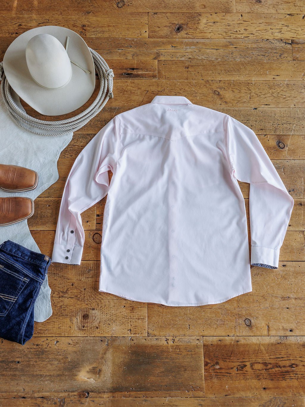 The Randall Ariat Men's Light Pink Button Up from Savannah Sevens western life{&}style is laid flat on a wooden floor beside a white cowboy hat, brown boots, and blue jeans.