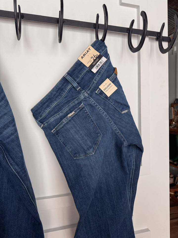 A pair of The Rawlins Perfect Rise Trouser Jeans by Savannah Sevens western life{&}style in medium wash hangs on a black metal wall hook, tags showing size 26R, against a white paneled wall.