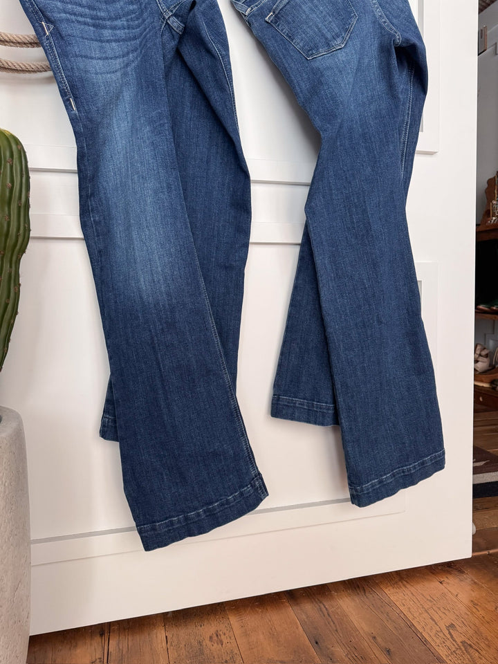 Two pairs of the Savannah Sevens western life{&}style The Rawlins Perfect Rise Trouser Jean *Exclusive* in medium wash hang side by side on a white door, with a green cactus in a gray pot and wooden floor in the foreground.