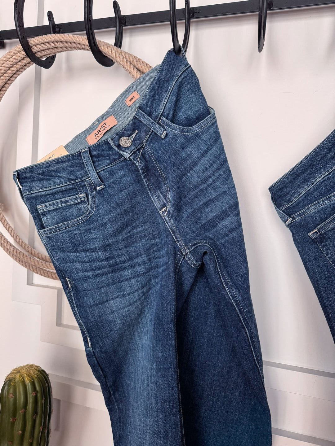 The Rawlins Perfect Rise Trouser Jean by Savannah Sevens western life{&}style in medium wash hangs on a black rack near a rope and small cactus, with another pair partially visible on the right.