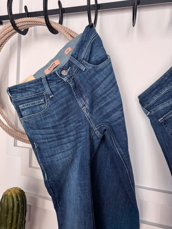 The Rawlins Perfect Rise Trouser Jean by Savannah Sevens western life{&}style in medium wash hangs on a black rack near a rope and small cactus, with another pair partially visible on the right.