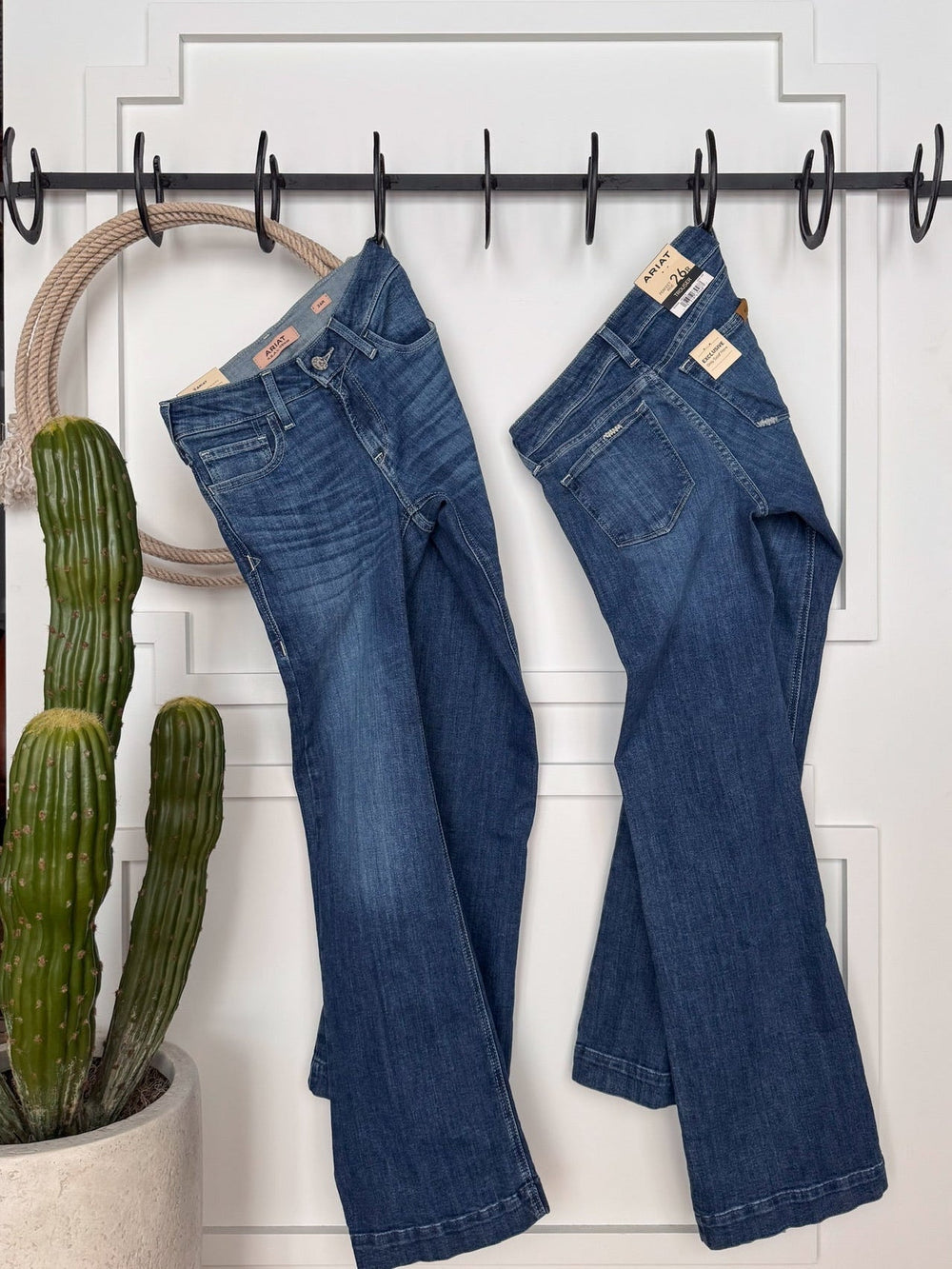 Two pairs of The Rawlins Perfect Rise Trouser Jean by Savannah Sevens western life{&}style hang on black hooks above a potted cactus, with a beige rope circle looped over one hook.