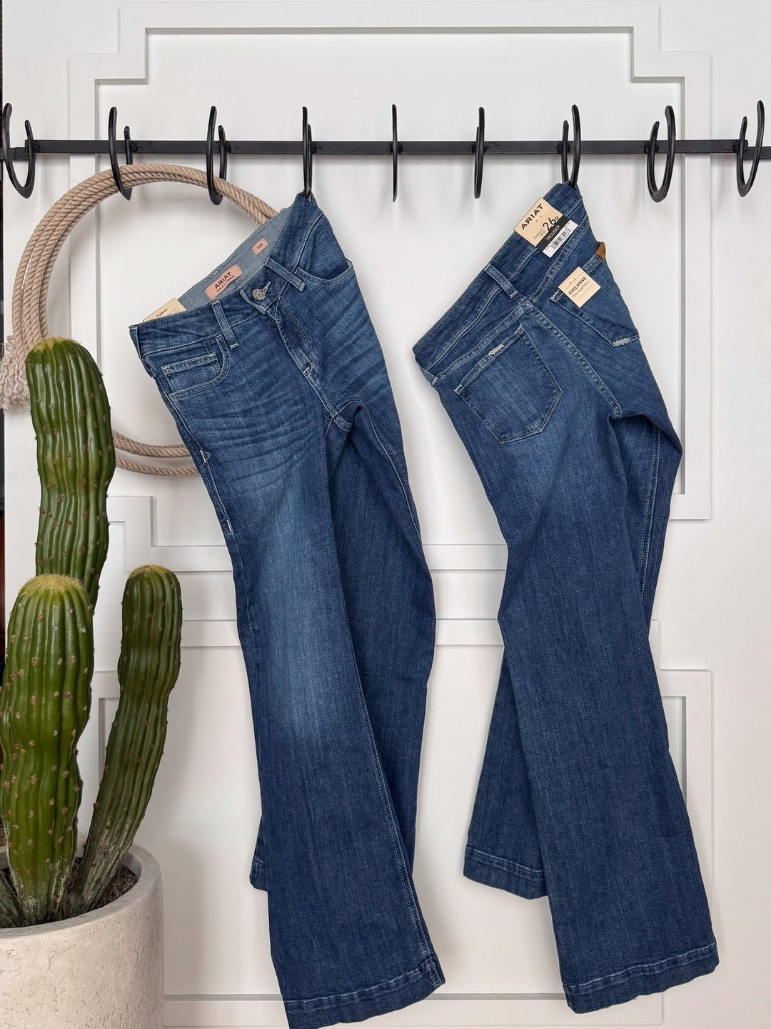 Two pairs of The Rawlins Perfect Rise Trouser Jean by Savannah Sevens western life{&}style hang on black hooks above a potted cactus, with a beige rope circle looped over one hook.