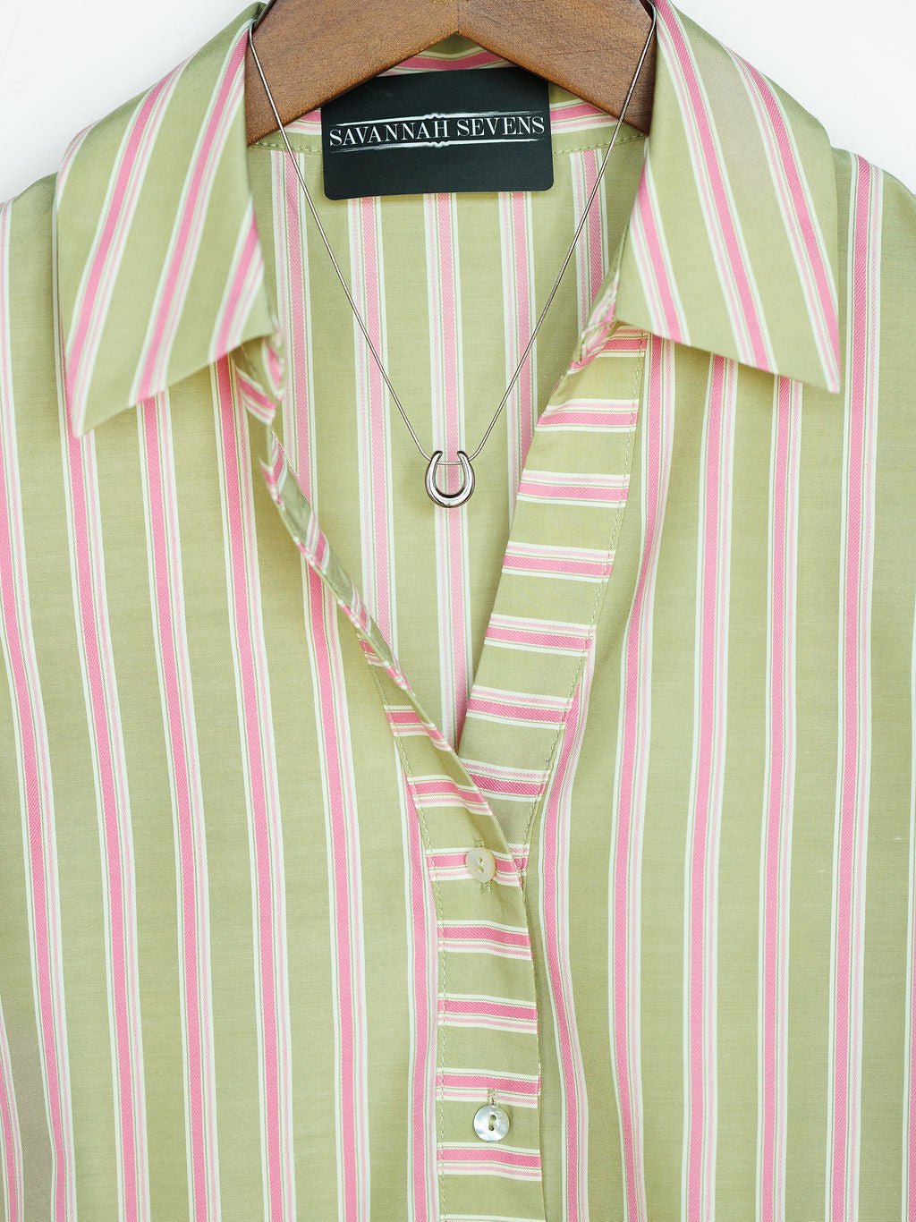 A close-up of The Rayas blouse by Savannah Sevens western life{&}style shows pink and white vertical stripes, buttoned up on a hanger, paired with a silver horseshoe pendant necklace.