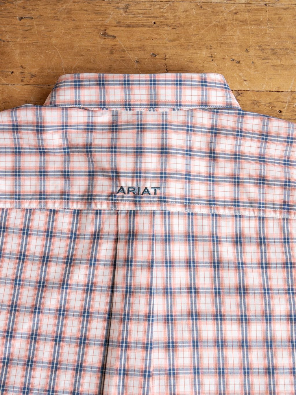 A close-up of the back of the Raymore Ariat Men's Plaid Button Up by Savannah Sevens western life{&}style, featuring peach, blue, and white plaid with ARIAT stitched above the yoke, displayed on a wooden surface.