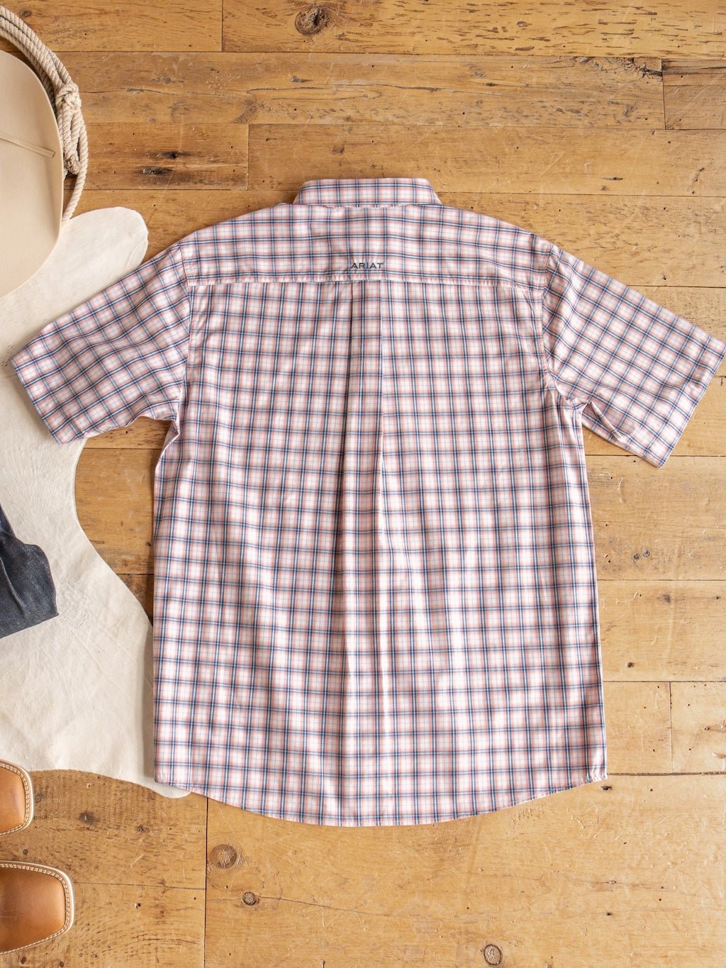 The Raymore Ariat Men's Plaid Button Up from Savannah Sevens western life{&}style is displayed laid flat on a wooden floor, showing the back, with other clothing items—including one from Ariat—partially visible at the edges.