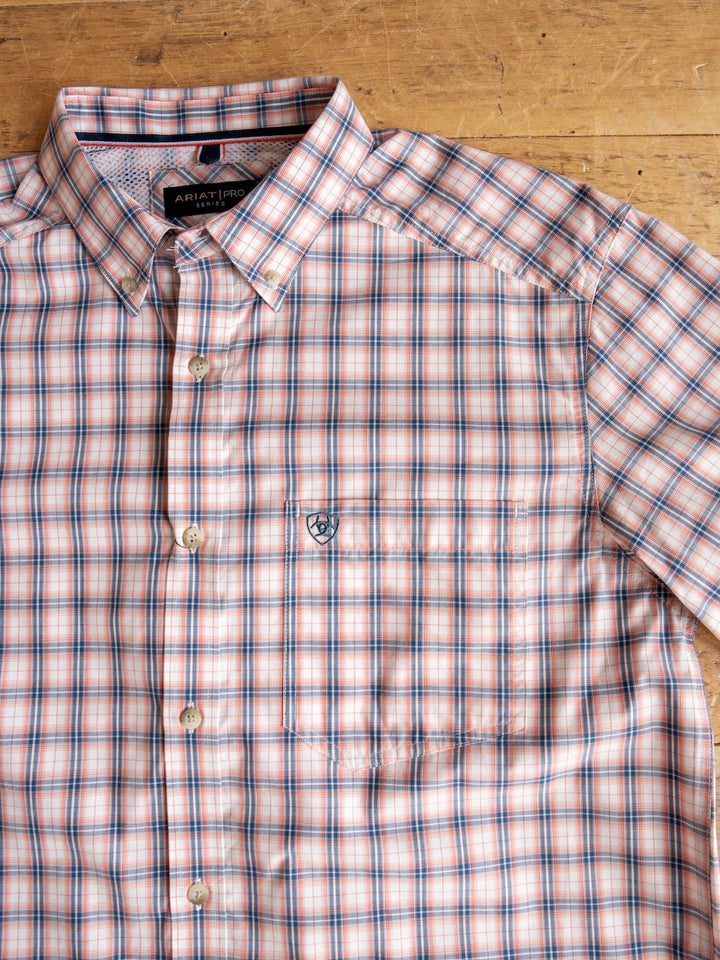 The Raymore Ariat Men's Plaid Button Up by Savannah Sevens western life{&}style is laid flat on a wooden surface. This short sleeve shirt features a red, white, and blue plaid pattern and has a front pocket adorned with a small embroidered logo.
