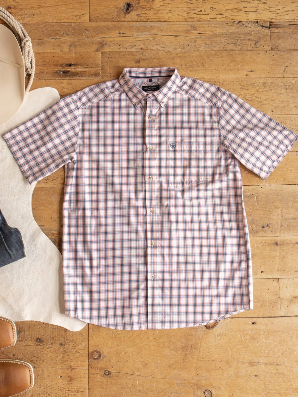 The Raymore Ariat Men's Plaid Button Up from Savannah Sevens western life{&}style, featuring a blue, brown, and white plaid pattern, is laid flat on a wooden floor alongside a hat, jeans, and tan shoes.