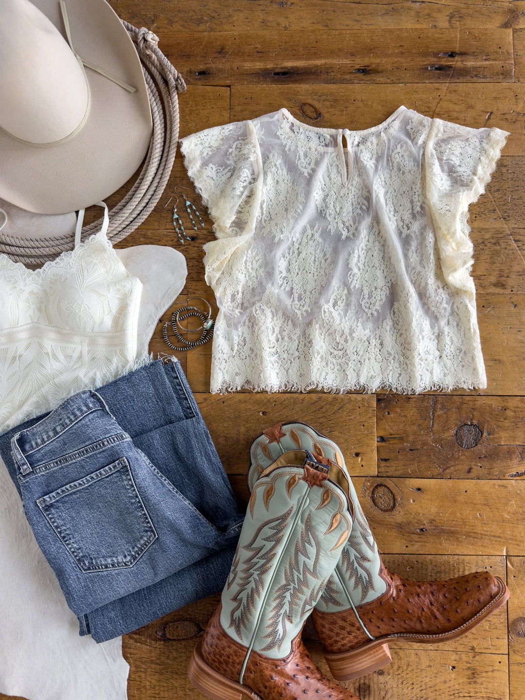 Flat lay of a Western outfit featuring a cream cowboy hat, Savannah Sevens western life{&}style Reata Cream Lace Top, white camisole, blue jeans, turquoise and brown cowboy boots, and silver hoop earrings arranged on a wooden surface.