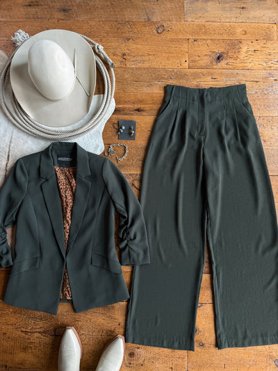 The Revillo Charcoal Olive Blazer Set from Savannah Sevens western life{&}style, featuring wide-leg dark green matte satin pants and a matching blazer, is styled with a brown-patterned top, a cream cowboy hat, cream boots, silver jewelry, and black earrings—all arranged on a wooden floor.