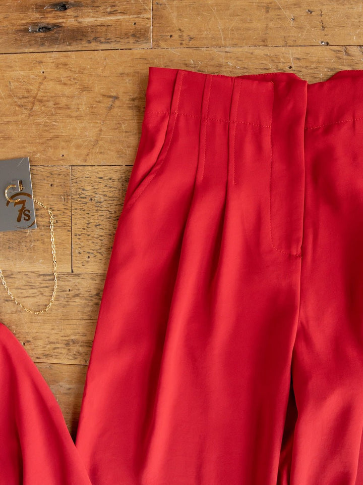 The Revillo Red Blazer Set from Savannah Sevens western life{&}style, featuring vibrant high-waisted pants with pleats and pockets, is displayed on a wooden surface. Beside the pants lies a gold chain necklace with a pendant and a tag labeled "7s.