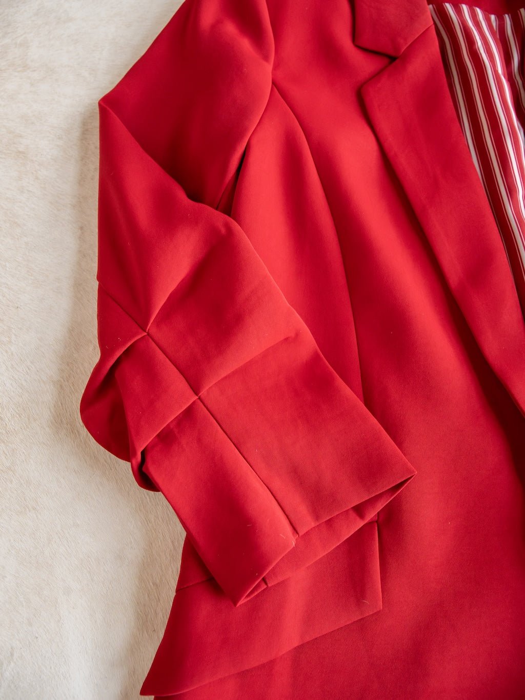 A close-up of the Revillo Red Blazer Set by Savannah Sevens western life{&}style shows the vibrant red satin blazer with folded sleeves resting on a light beige surface. The blazer, which is part of a coordinated set, features a striped lining visible at the collar.