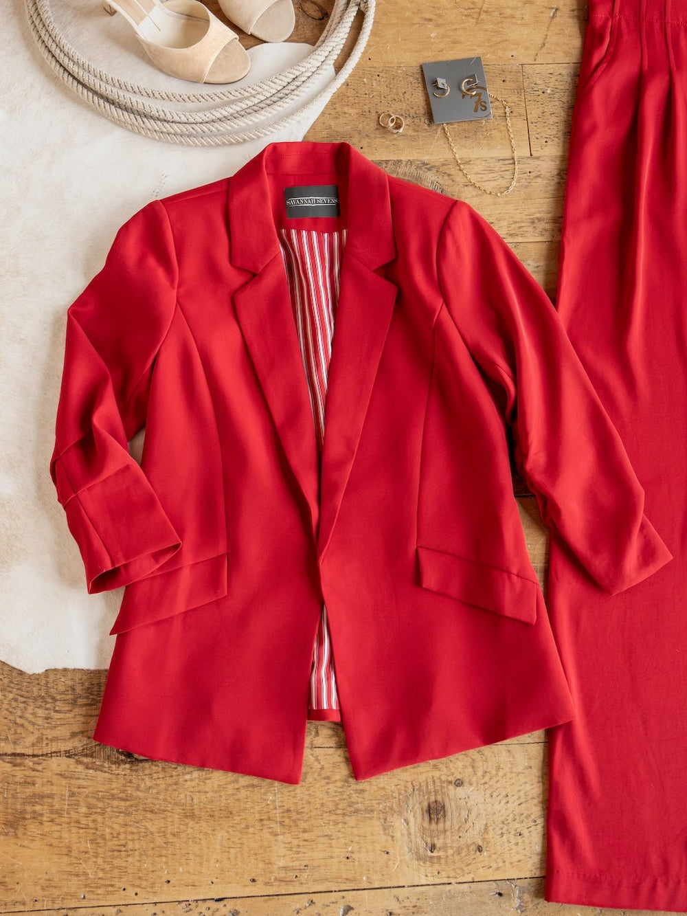 The Revillo Red Blazer Set by Savannah Sevens western life{&}style, featuring a red satin blazer with striped lining, is laid flat on a wooden surface alongside the matching high waisted pants, beige sandals, and gold jewelry.