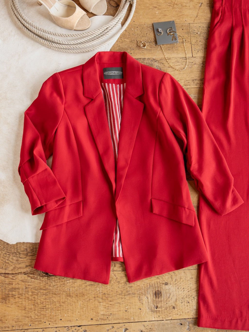 The Revillo Red Blazer Set by Savannah Sevens western life{&}style, featuring a red satin blazer with striped lining, is laid flat on a wooden surface alongside the matching high waisted pants, beige sandals, and gold jewelry.